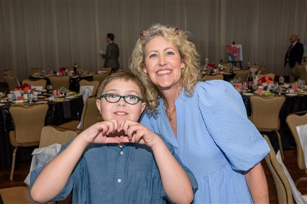 An artist and a student smile at the camera. The student makes a heart shape with his hands while the artist smiles wide. Both are happy and joyful. 